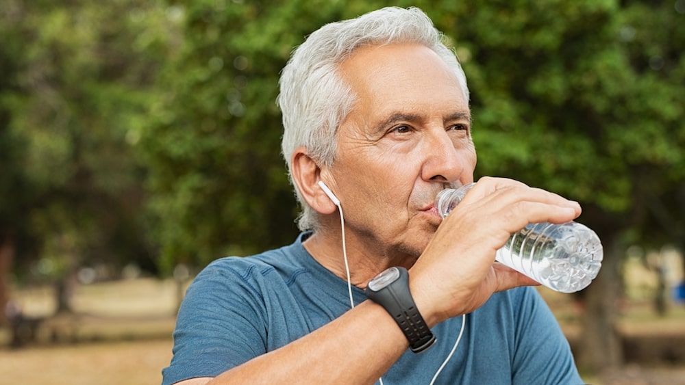 En äldre man med grått hår och blå t-shirt dricker vatten ur en flaska.