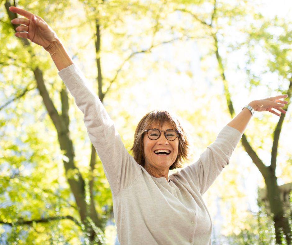 En kvinna på ca 60 år räcker upp händerna i luften och ler i en skogsglänta.