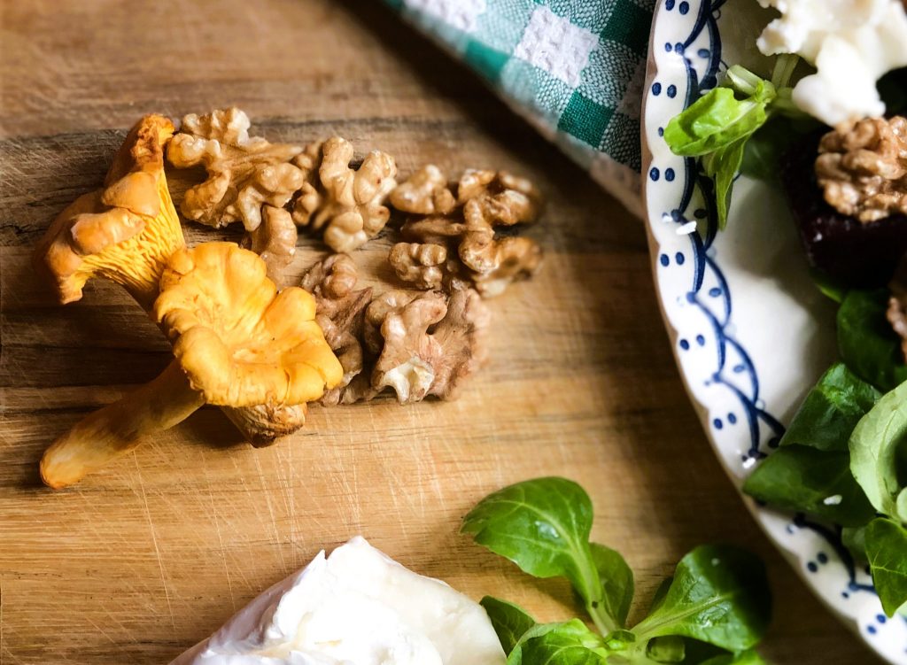 Walnuts and chanterelle mushrooms on a wooden chopping board next to a blue and white plate with salad on top