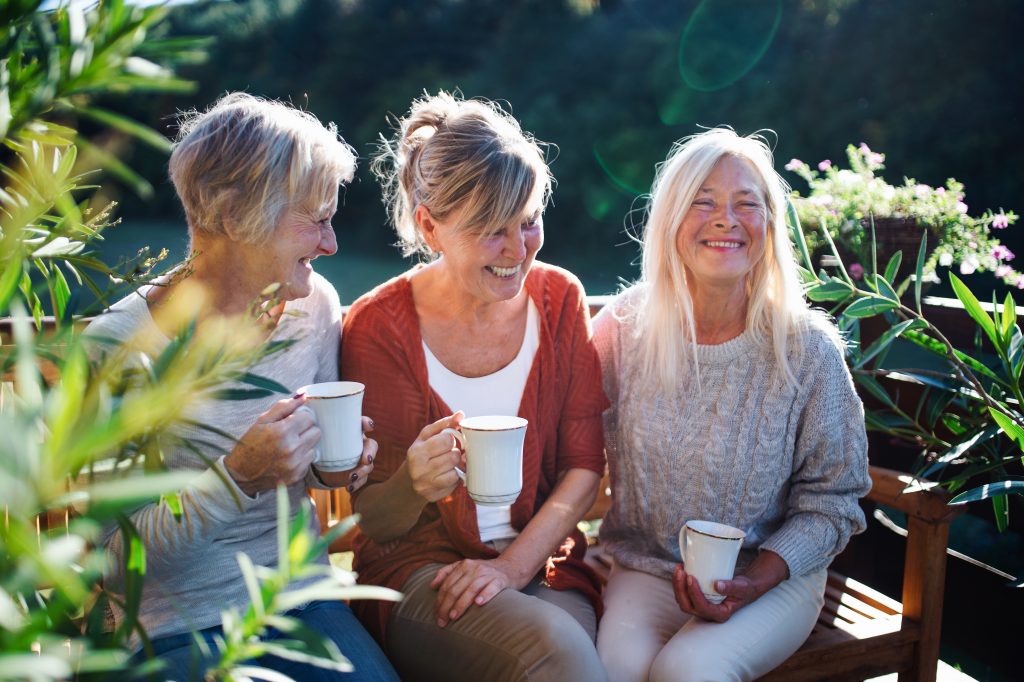 Tre kvinnor i 60-årsåldern sitter på en bänk i solen och dricker kaffe och skrattar. 
