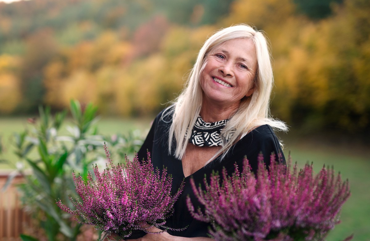 A woman standing outdoors on terrace holding heather flowers, looking at the camera.
