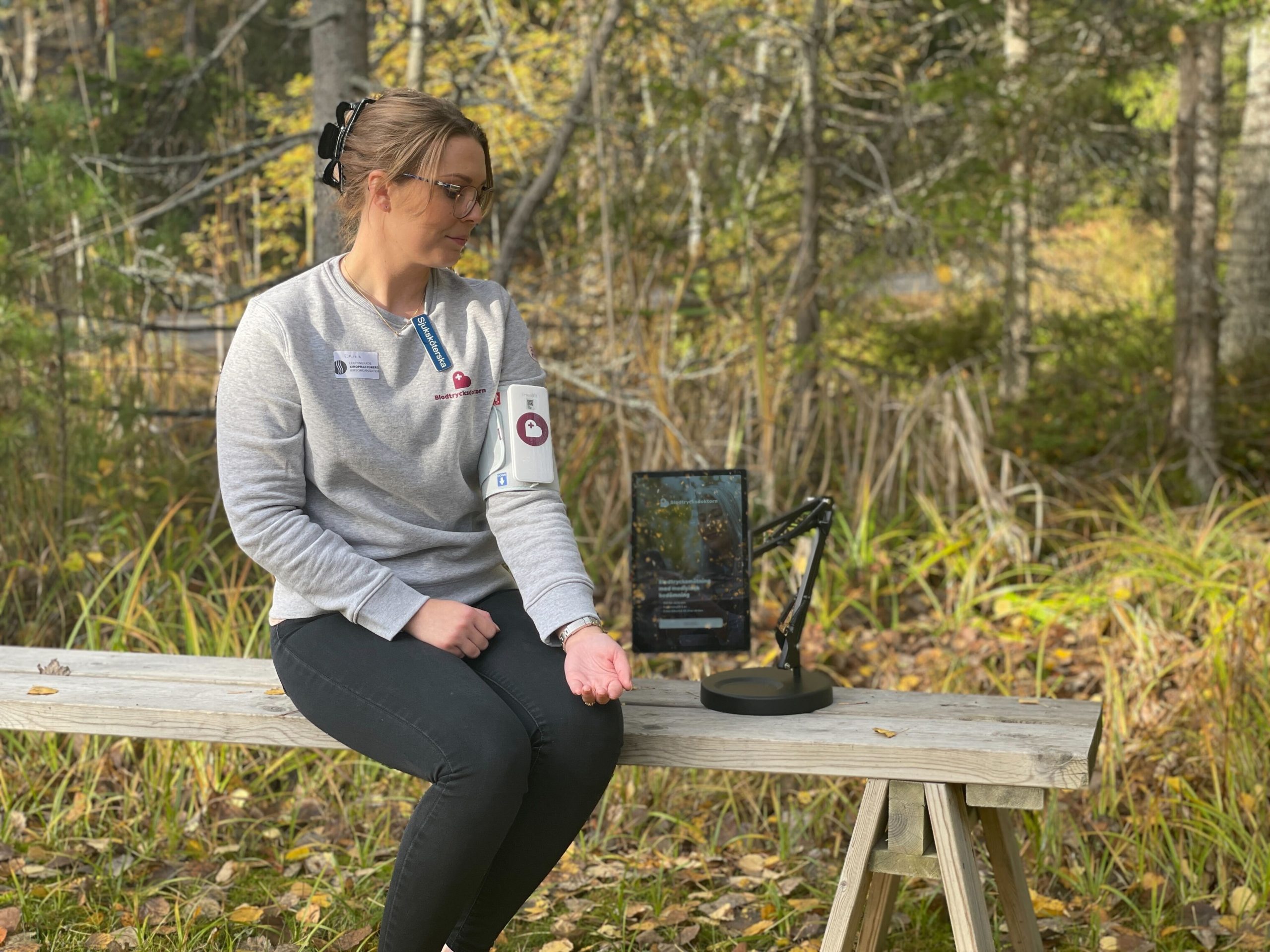 Sjuksköterskan Erika Mankenskiöld sitter på en bänk i skogen, hon mäter sitt blodtryck med en blodtrycksmanschett och en surfplatta.