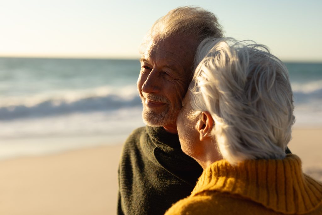 Ett äldre par står nära varandra och ler på en strand. 