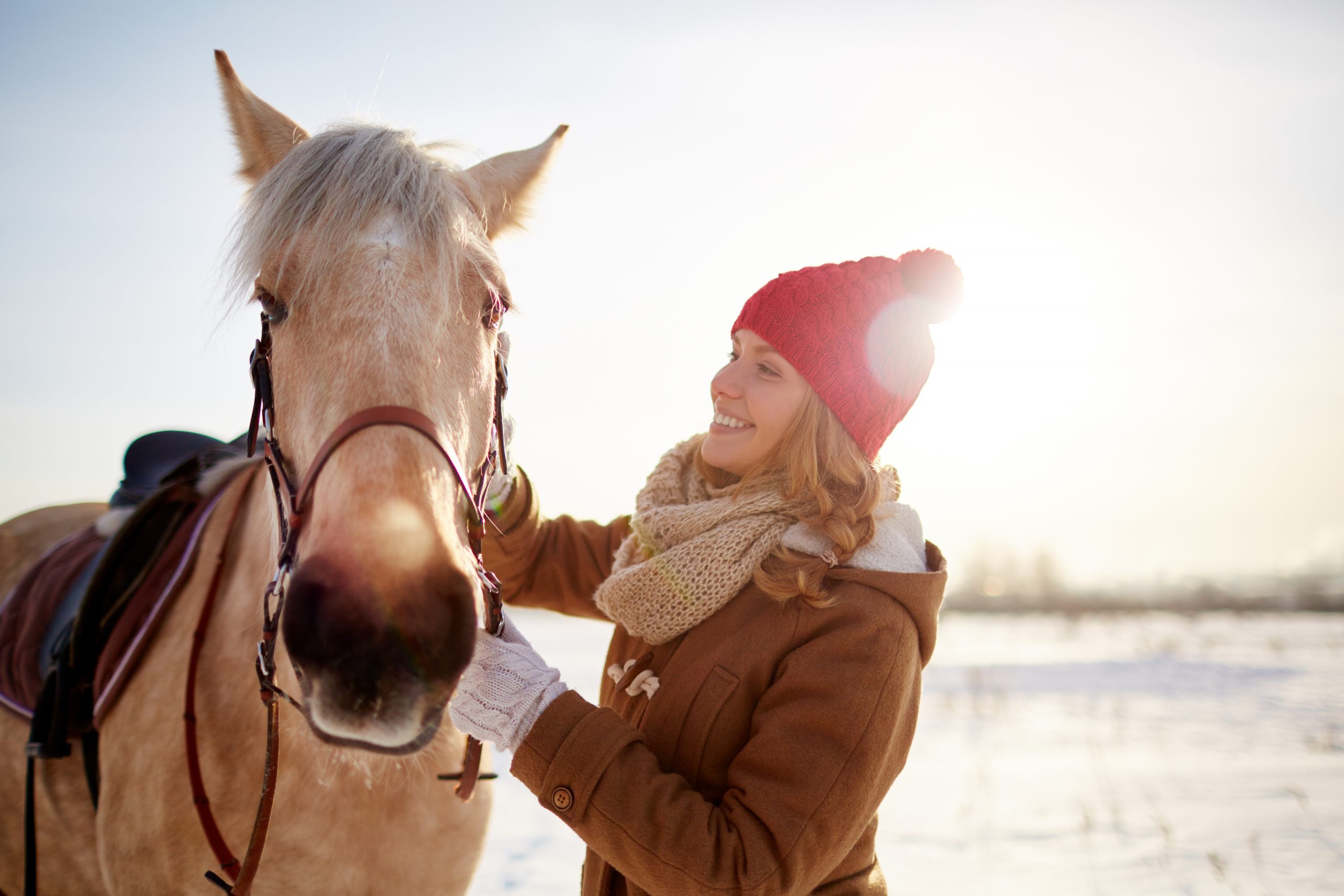 En blond kvinna med vinterkläder klappar en beige häst ute i snön.
