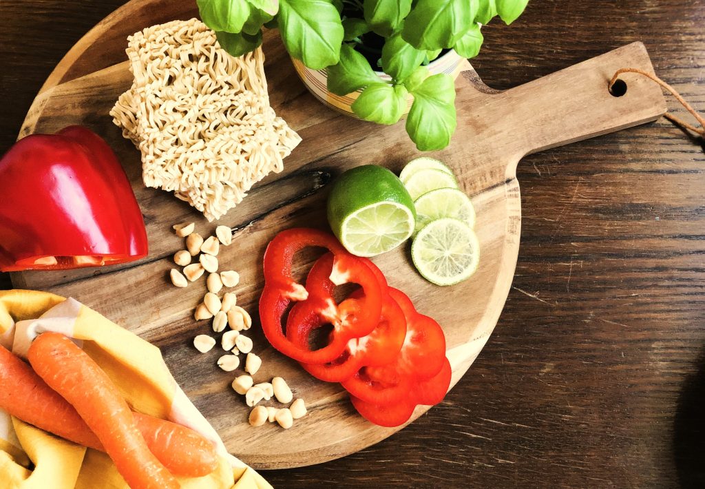 Egg noodles, basil, carrots, peanuts, red bell peppers and a sliced lime placed on a round wooden chopping board