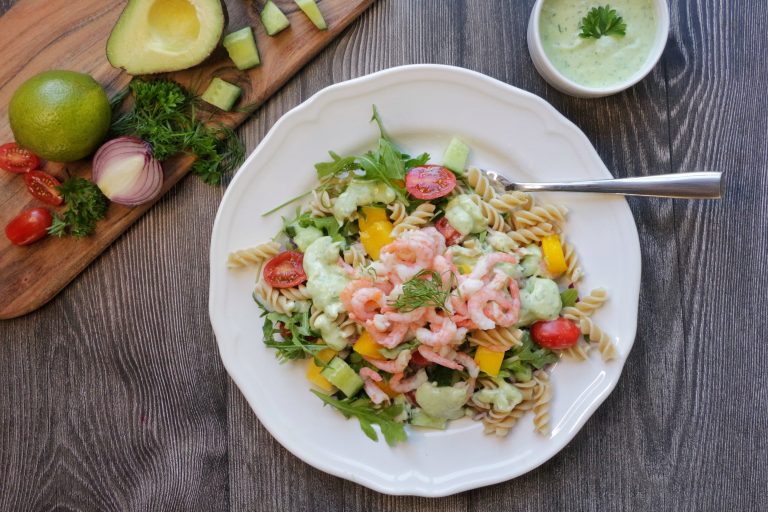 Pastasallad med räkor och grönsaker på en vit tallrik, ståendes på ett grått träbord. Bredvid finns en skärbräda med grönsaker. 