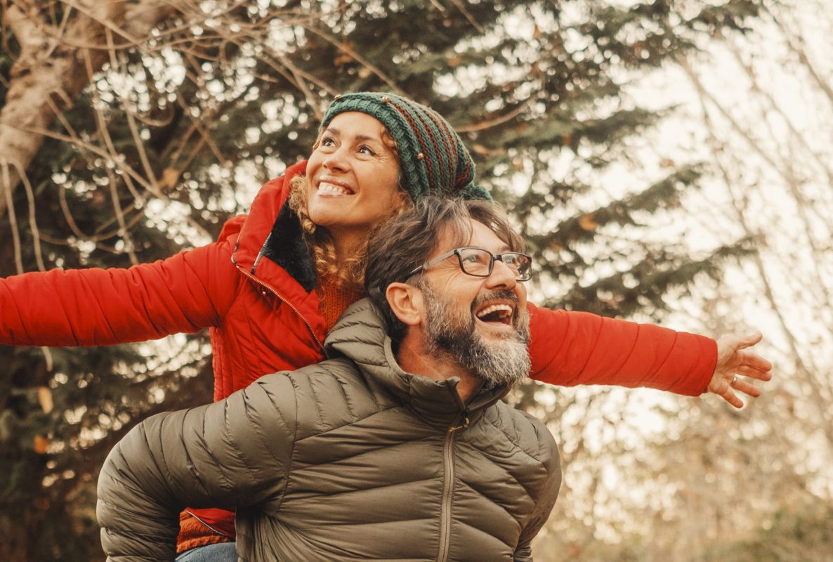 Medelålders man bär en kvinna på ryggen ute i naturen, båda är klädda i vinterkläder och skrattar