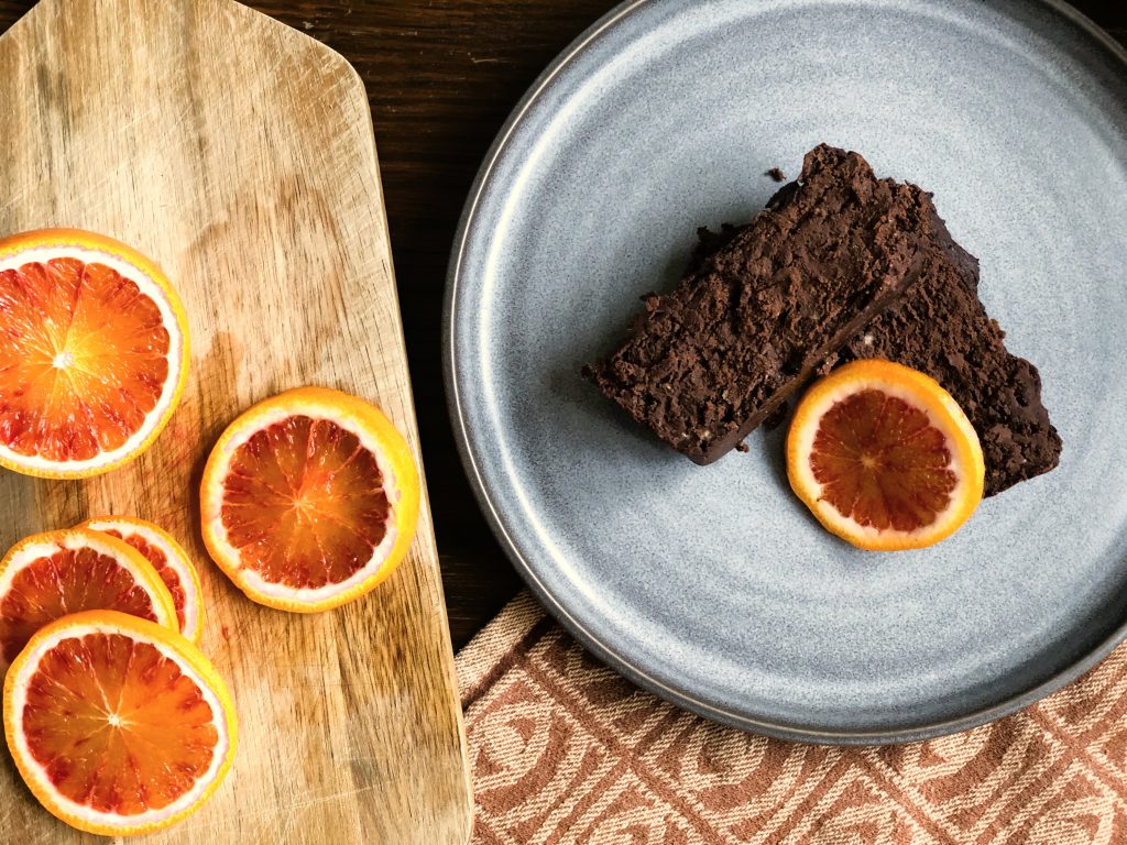 Två bitar brownies liggandes på en grå assiett, bredvid finns en skärbräda med apelsinskivor. 