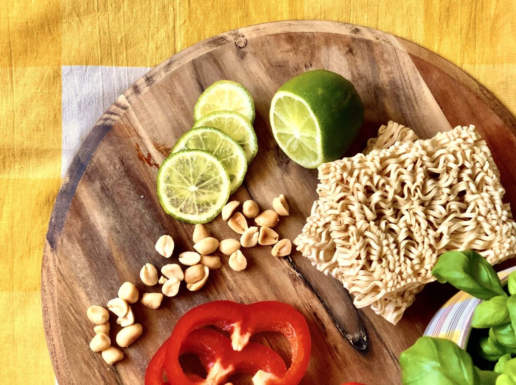 Egg noodles, peanuts, red bell peppers and a sliced lime placed on a round wooden chopping board