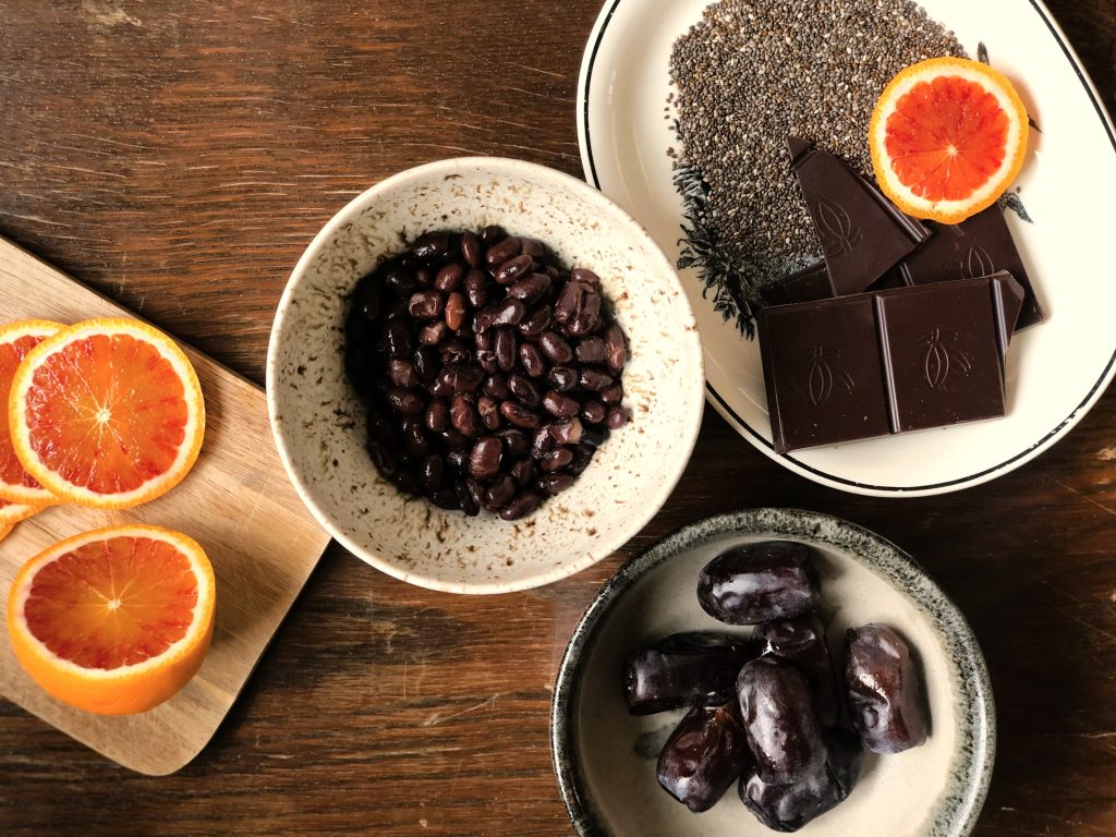Dadlar i en skål, apelsin på ett skärbräde, mörk choklad i en skål och svarta bönor i en skål. 