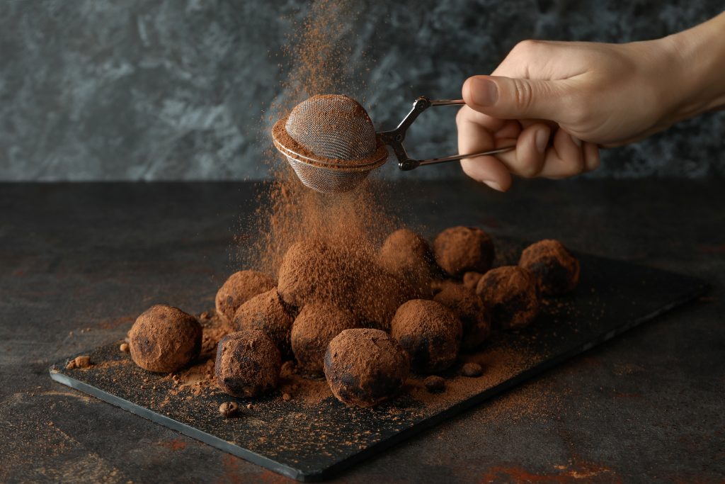 En hand som strör kakaopulver över chokladtryfflar med en tesil. 