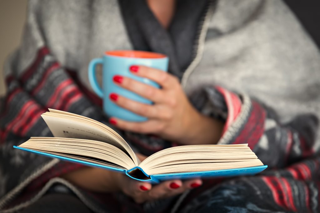 En kvinna läser en bok och dricker te med en filt runt sig. 