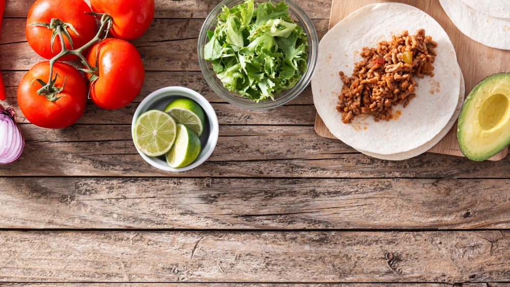 Ingredienser till tacos, rödlök, tomat, lime, sallad, tortillabröd, färs och avokado, liggandes på ett träbord