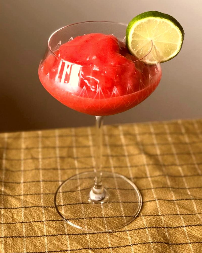 En röd jordgubbsdrink med en limeskiva i ett glas på fot. 