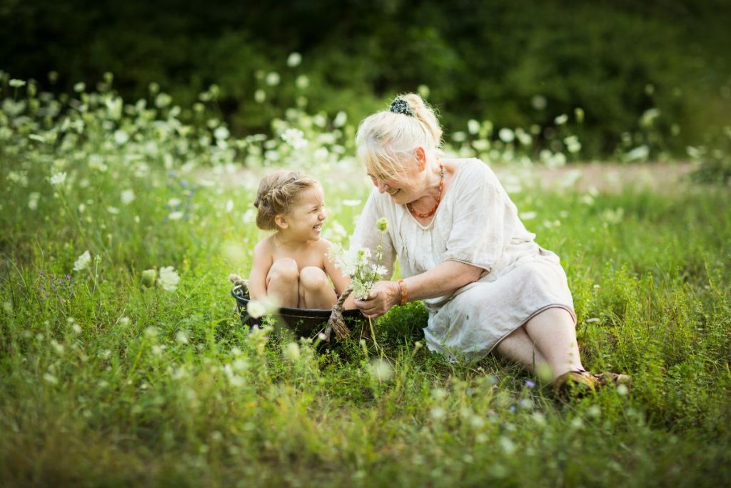 En äldre kvinna i beige klänning bara en liten flicka på ca 2 år i en badbalja på en blommig äng.