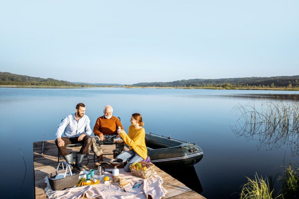 Ung man och kvinna har picknick med en gammal man på en solig brygga vid vattnet.
