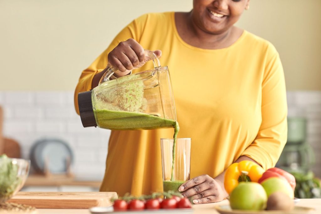 Leende kvinna i gul tröja häller upp en grön smoothie i ett glas.