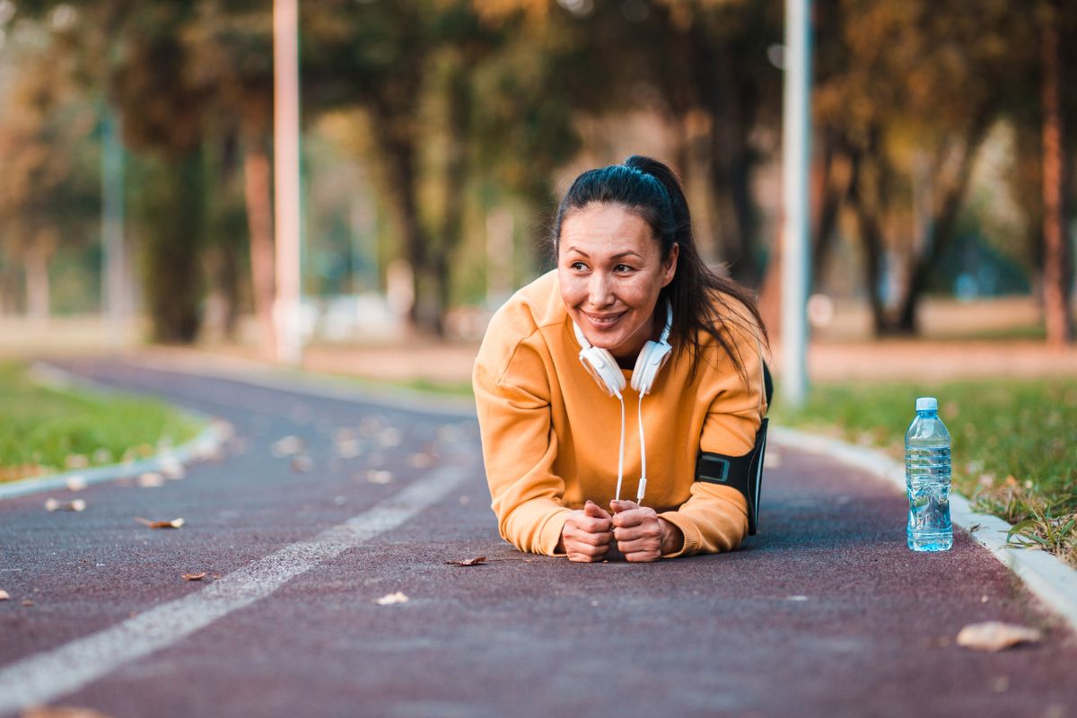 Glad kvinna med mörkt hår och orange jacka står i plankan på en löparbana