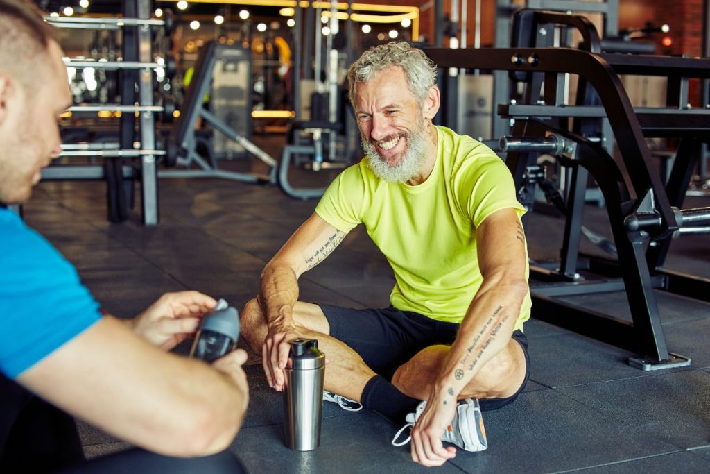 Två medelålders män pratar och skrattar på golvet i ett gym