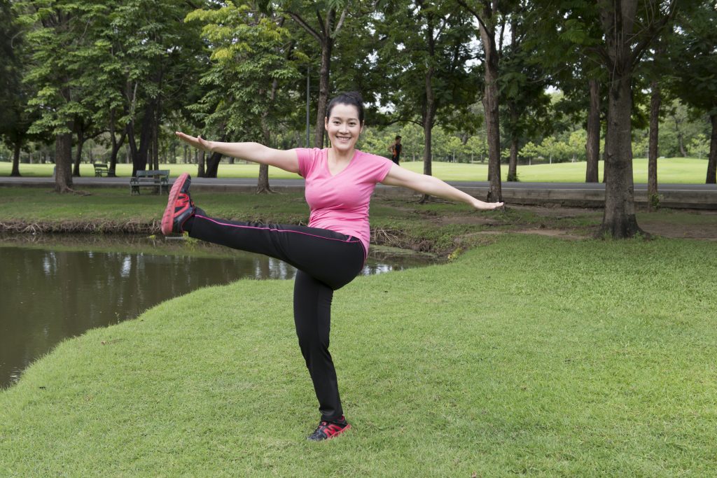 Kvinna ca 30 år står på ett ben och balanserar utomhus i en park. Hon bär svarta byxor och en rosa träningströja.