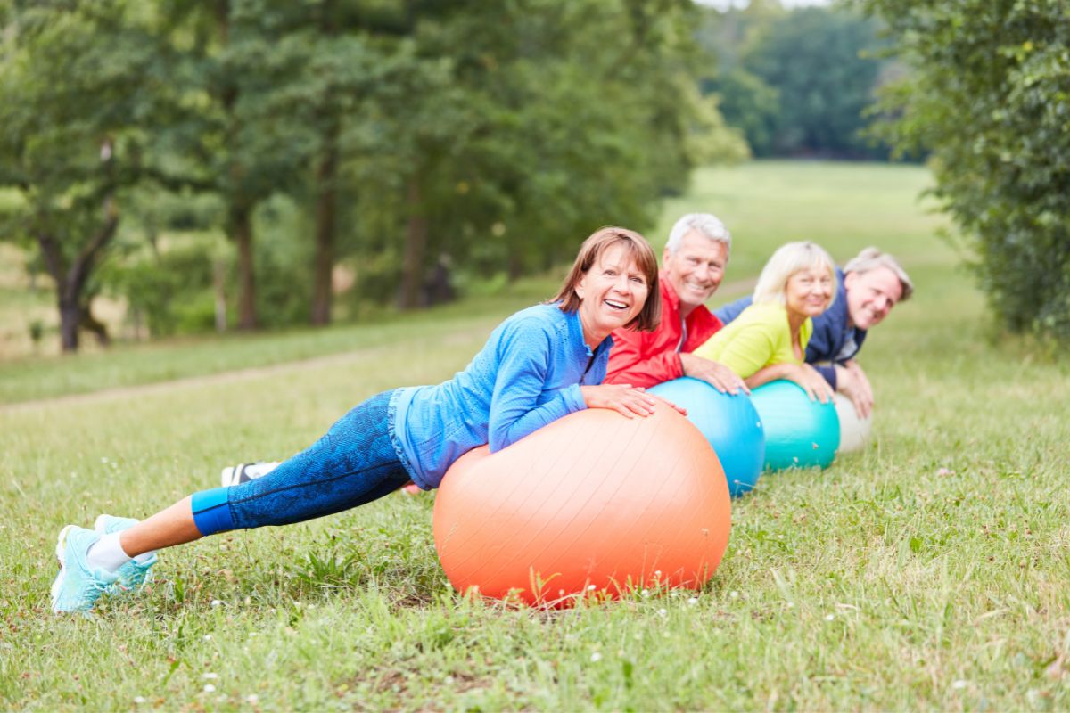 grupp seniorer står i plankan på pilatesbollar på en gräsplätt