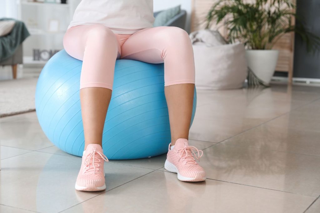 kvinna i rosa träningsbyxor och sneakers sitter på en blå pilatesboll
