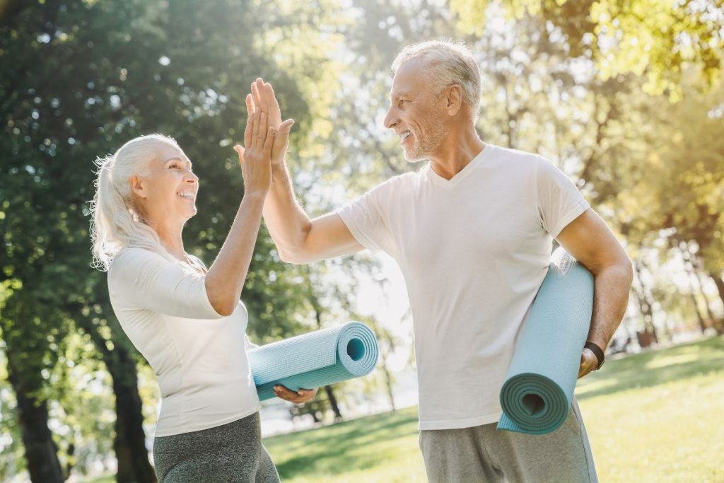 En stilig man och kvinna i vita tröjor ger varandra en high five efter ett yogapass i en solig park
