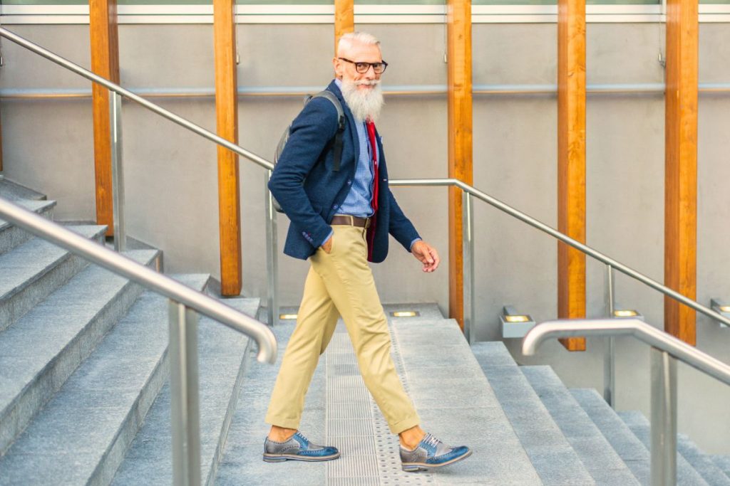 Vithårig man med skägg och glasögon, ca 60 år, iklädd blå kavaj och beiga chinos, går nerför en trappa