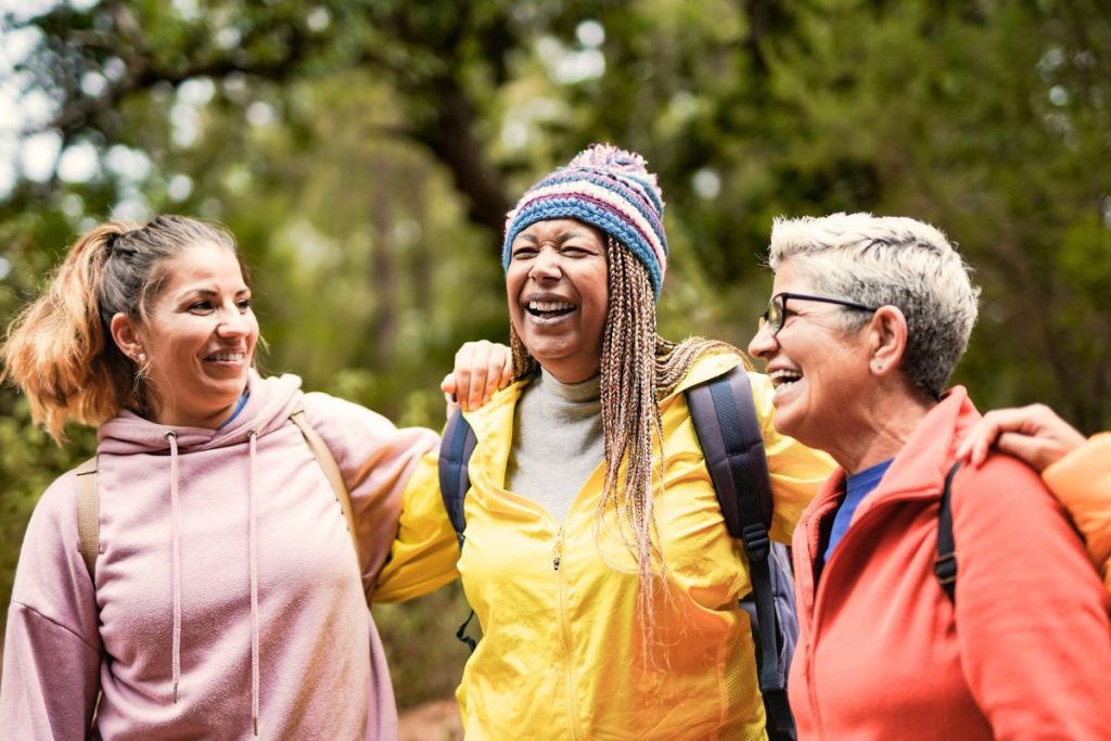 Tre kvinnor rmellan 35 och 45 år,  iklädda färgglada tröjor, håller om varandra och skrattar under en promenad i skogen.