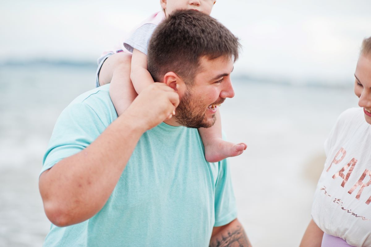 En man, ca 30 år, med brunt hår och ljusblå t-shirt bär ett litet barn på axlarna på en strand