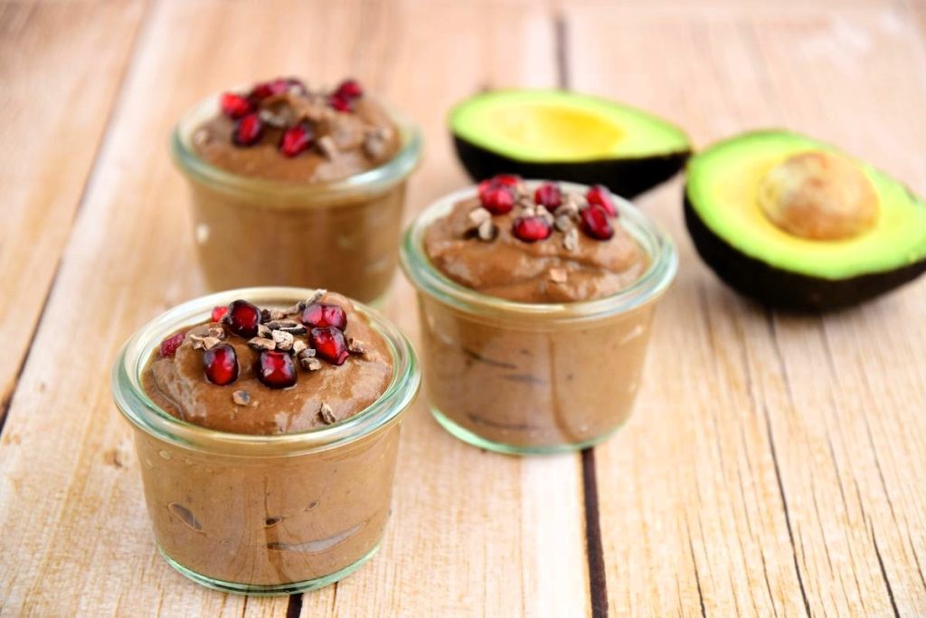 Tre glasbägare med chokladmousse och granatäppelkärnor ståendes på ett träbord. I bakgrunden ligger en delad avokado. 