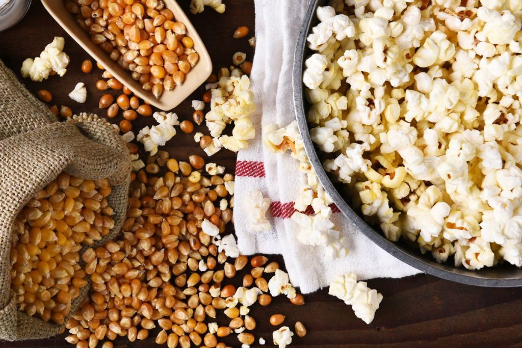 En kastrull med popcorn på ett träbord, bredvid ligger en påse och en skopa med popcornkärnor