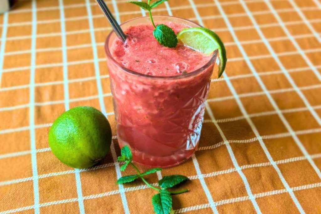 En frusen drink gjord på vattenmelon, lime och mynta står på ett bord med en orangerutig duk. Bredvid ligger en lime och en myntakvist.