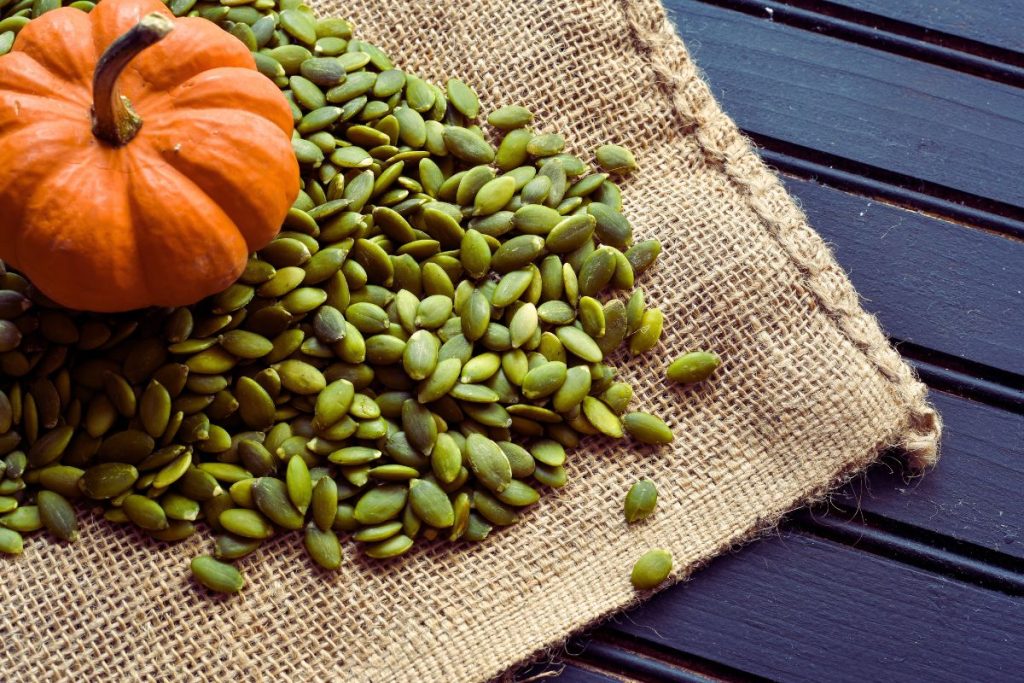 En hög pumpakärnor och en liten halloweenpumpa ligger på en juteduk på ett bord.
