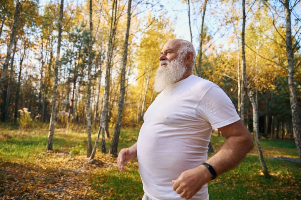 Äldre man med vitt skägg och vit t-shirt joggar i en skog på hösten.