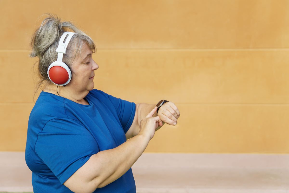 Kraftig kvinna iklädd blå träningströja och hörlurar, ca 55 år, kollar på sin smartklocka under ett träningspass.