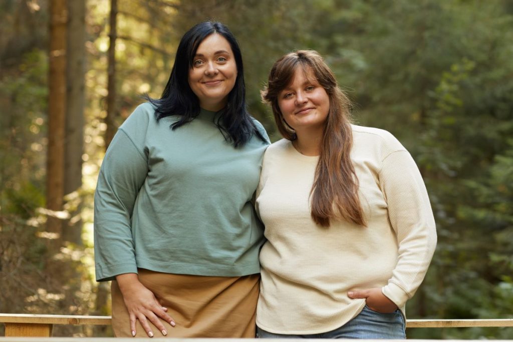 Två kurviga kvinnor, ca 30 år gamla, håller om varandra och ler mot kameran på en bro i en skog.