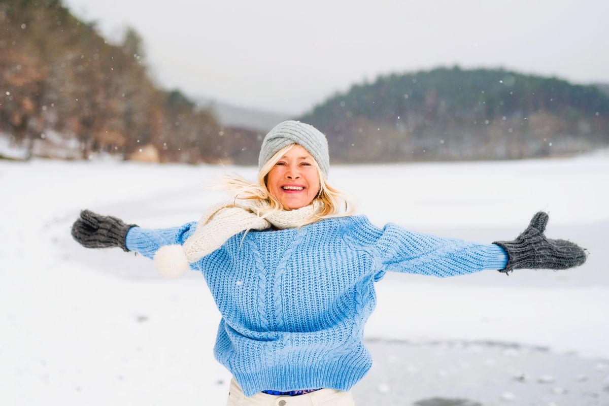 Glad kvinna, ca 55 år, ler och håller ut armarna vid en frusen sjö.