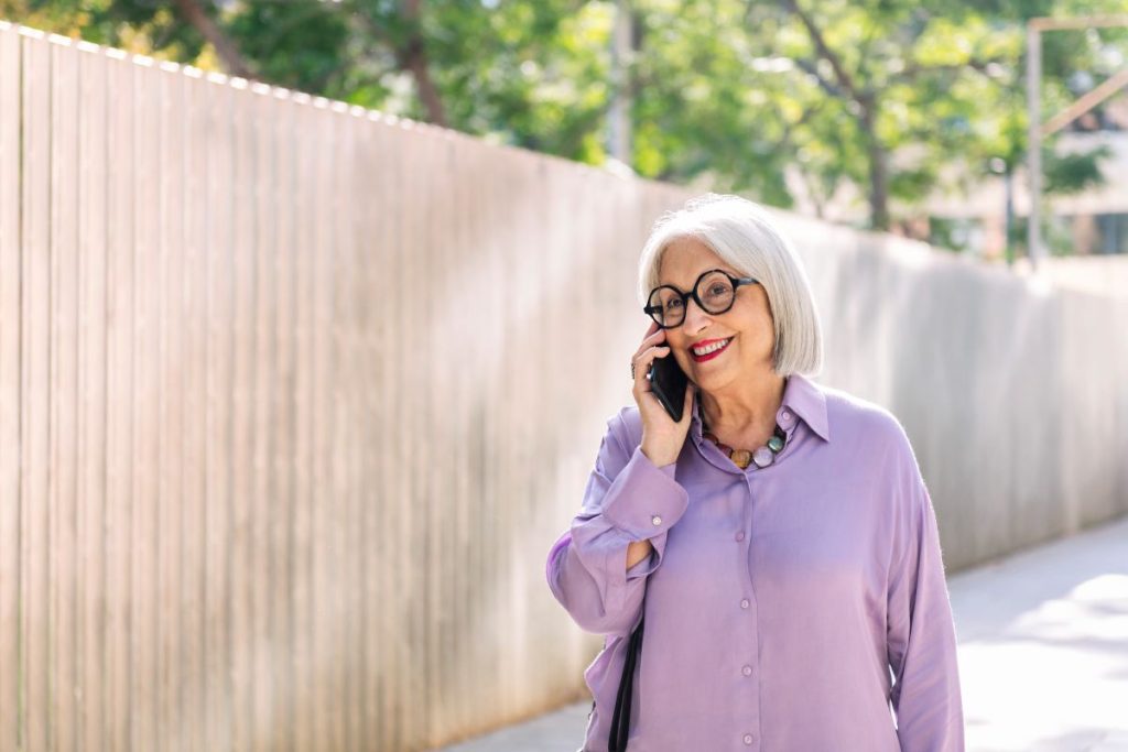 Vacker kvinna ca 65 år gammal i lila skjorta tar en promenad i solen och pratar i telefon