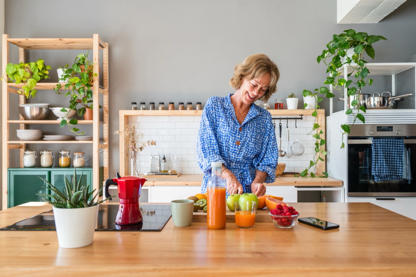 En kvinna i blå skjorta står i sitt kök med frukt och grönsaker på bänken