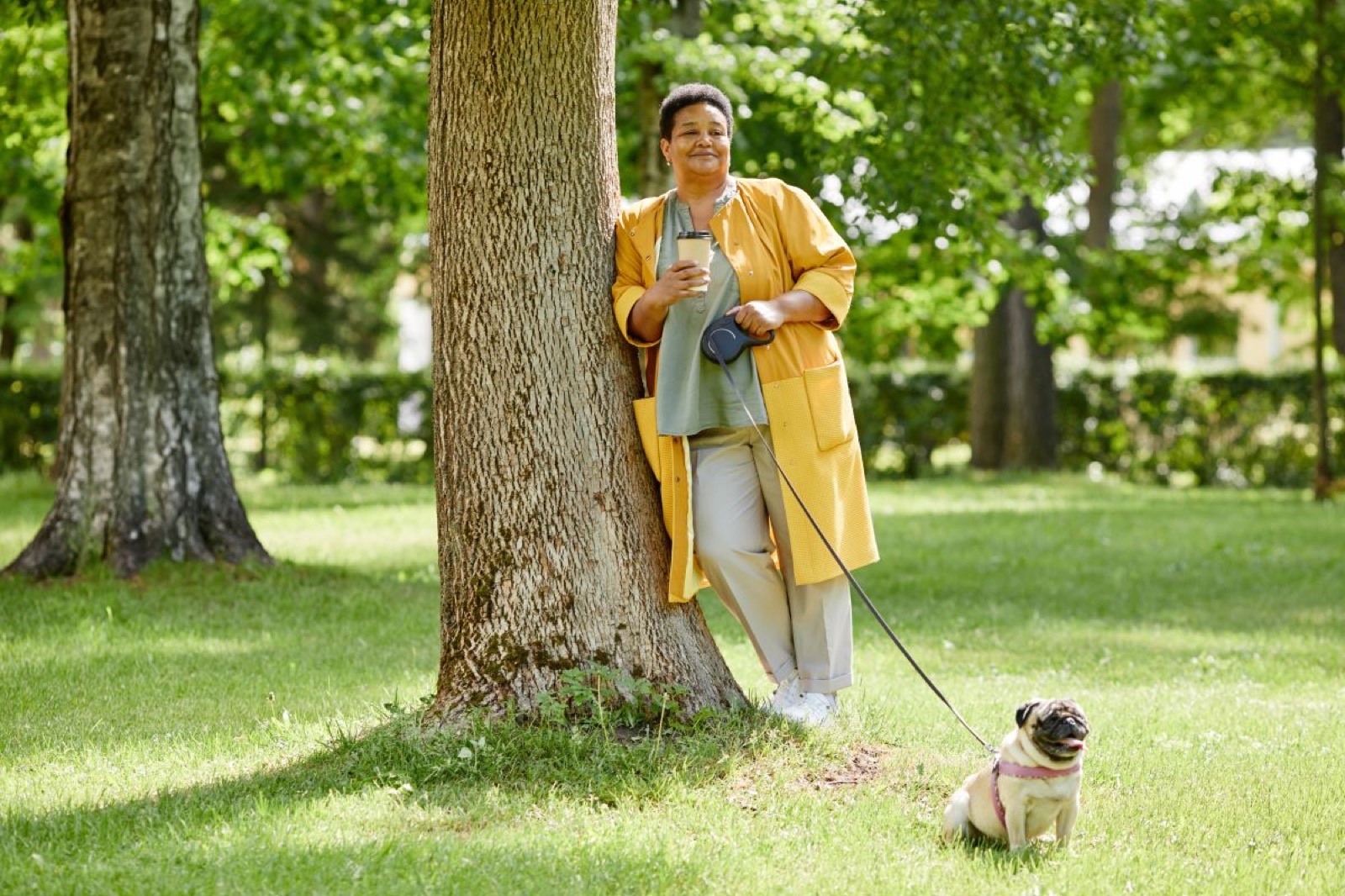 En kvinna i gul kappa lutar sig mot ett träd i en park med en hund i koppel